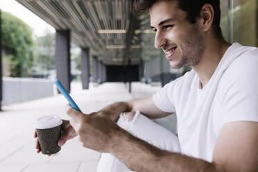 Smiling young man with disposable cup using mobile phone at corridor - XLGF02087