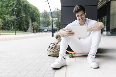 Mann mit Kopfhörern und digitalem Tablet auf dem Skateboard an der Säule - XLGF02090