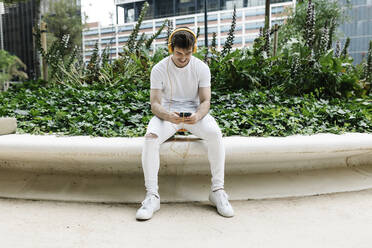 Man wearing headphones using mobile phone on retaining wall - XLGF02097