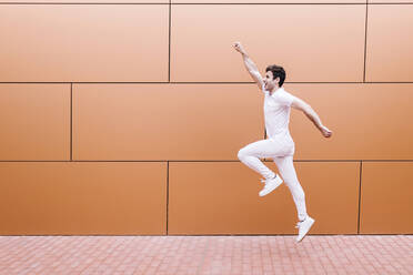 Cheerful young man jumping on footpath by orange wall - XLGF02100