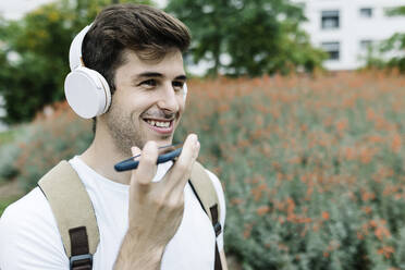 Lächelnder junger Mann, der eine Sprachnachricht über ein Mobiltelefon an eine Wiese sendet - XLGF02111