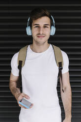 Young man holding mobile phone while listening music through headphones in front of shutter - XLGF02114