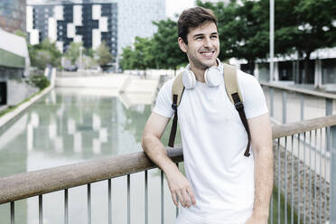Lächelnder junger Mann mit Rucksack, der den Blick abwendet, während er sich auf ein Geländer am See stützt - XLGF02117