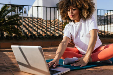 Fokussierte afroamerikanische Frau mit lockigem Haar, die ein Online-Tutorial am Laptop auswählt, während sie auf einer Matte auf dem Dach sitzt und sich auf eine Yogastunde vorbereitet - ADSF25353