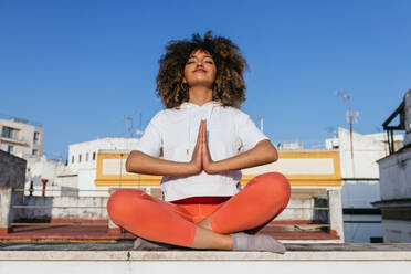 Ruhige afroamerikanische Frau sitzt in Padmasana mit Namaste-Geste und meditiert mit geschlossenen Augen, während sie auf einem Dach Yoga übt - ADSF25361