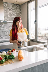 Young homosexual female pouring water from tap into saucepan with uncooked pasta against assorted vegetables in house - ADSF25378