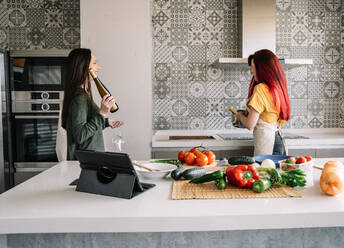 Content young homosexual girlfriends with bottle of wine speaking while looking at each other between stove and fresh vegetables - ADSF25379