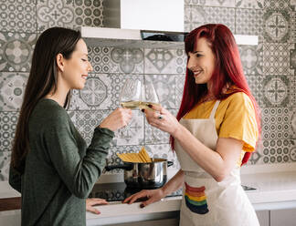 Seitenansicht von jungen lächelnden homosexuellen Frauen mit Gläsern und einer Flasche Weißwein, die sich im Haus unterhalten, während sie sich gegenseitig ansehen - ADSF25382