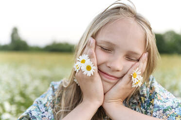 Blond girl with chamomile flowers smiling while day dreaming in field - EYAF01695