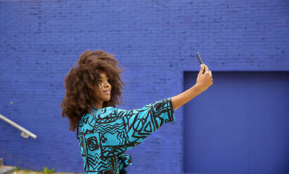 Young woman taking selfie through smart phone in front of blue wall - FMKF07271