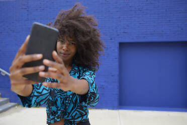 Woman taking selfie through smart phone in front of blue wall - FMKF07272