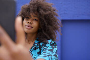 Afro woman taking selfie through mobile phone in front of blue wall - FMKF07274