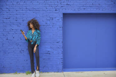 Afro-Frau hält Handy und lehnt sich an eine blaue Backsteinmauer - FMKF07276