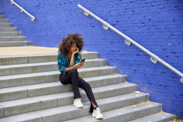 Junge Frau benutzt ihr Smartphone, während sie auf einer Treppe sitzt - FMKF07279