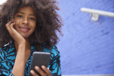 Lächelnde Afro-Frau mit Hand am Kinn, die ein Smartphone hält - FMKF07280