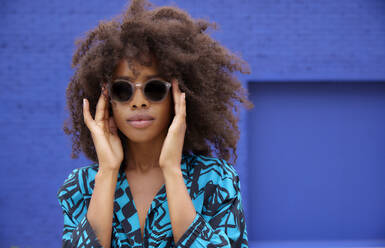 Afro woman wearing sunglasses in front of blue wall - FMKF07290