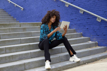 Afro-Frau benutzt digitales Tablet, während sie auf einer Treppe sitzt - FMKF07294