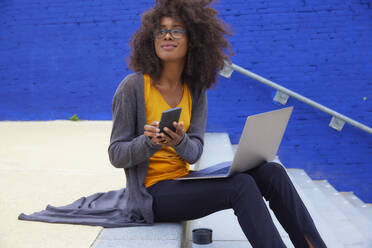 Afro-Frau sitzt mit Handy und Laptop auf einer Treppe - FMKF07300