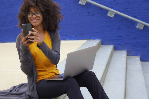Lächelnde Afro-Frau, die ein Mobiltelefon benutzt, während sie auf einer Treppe sitzt - FMKF07302