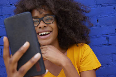 Smiling woman taking selfie through mobile phone in front of brick wall - FMKF07305