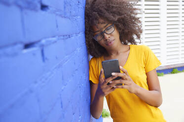 Afro woman using mobile phone while leaning on blue wall - FMKF07306