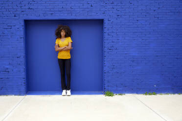 Afro-Frau mit verschränkten Armen auf dem Fußweg vor einer blauen Backsteinmauer stehend - FMKF07312