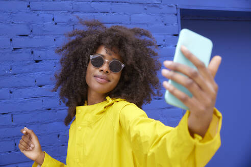 Young woman wearing sunglasses taking selfie through smart phone in front of blue wall - FMKF07325