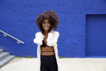 Lächelnde schöne Afro-Frau, die vor einer blauen Wand wegschaut - FMKF07333