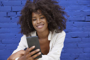 Lächelnde Afro-Frau, die ein Smartphone vor einer blauen Wand benutzt - FMKF07341