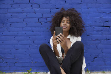 Junge Afro-Frau, die mit ihrem Mobiltelefon vor einer blauen Wand im Internet surft - FMKF07342