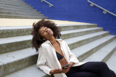 Junge Afro-Frau mit geschlossenen Augen, die auf einer Treppe sitzend Musik über In-Ear-Kopfhörer hört - FMKF07344