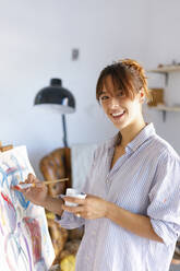 Smiling female painter holding paintbrush while painting in studio - IFRF00902