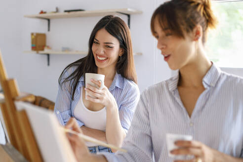 Glückliche Künstlerinnen, die eine Tasse in der Hand halten, während ein Künstlerkollege im Atelier malt - IFRF00911