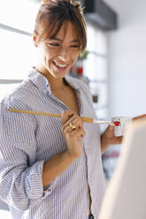 Smiling young female artist holding paintbrush looking at painting in studio - IFRF00915