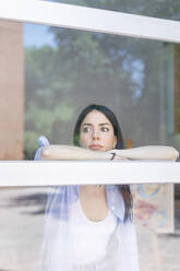 Weibliche Fotografin, die wegschaut, während sie sich im Studio an ein Fenster lehnt, gesehen durch Glas - IFRF00926