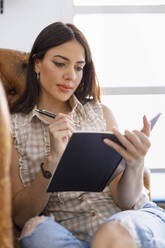 Woman writing in notebook while sitting on armchair at art studio - IFRF00943