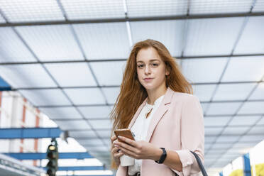 Redhead businesswoman holding mobile phone and disposable coffee cup under shade on railway platform - EIF01449