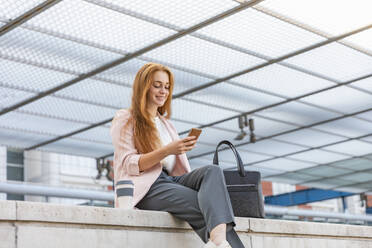 Young female professional smiling while using mobile phone on retaining wall at railway station - EIF01457