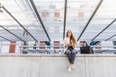 Freiberuflerin mit Mobiltelefon, die auf einer Stützmauer am Bahnhof sitzt und wegschaut - EIF01461