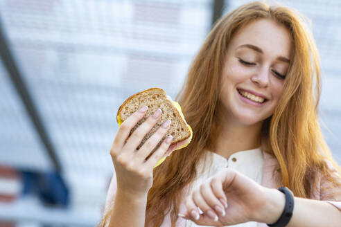 Lächelnde weibliche Fachkraft, die die Zeit überprüft, während sie ein Sandwich am Bahnhof isst - EIF01466