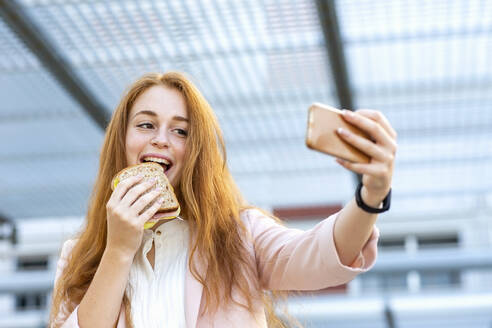 Rothaarige Geschäftsfrau nimmt Selfie durch Handy beim Essen Sandwich - EIF01467