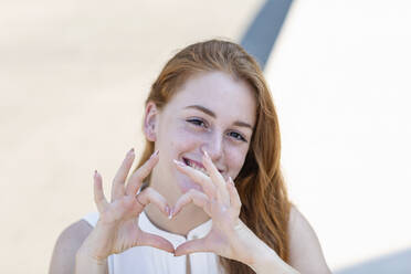 Smiling businesswoman with redhead showing heart shape gesture during sunny day - EIF01479
