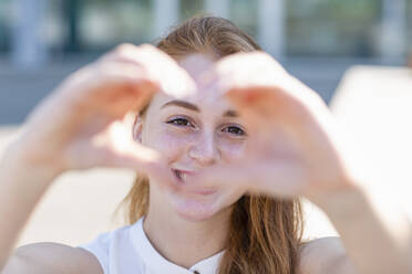 Happy female freelancer looking through heart shape gesture during sunny day - EIF01480