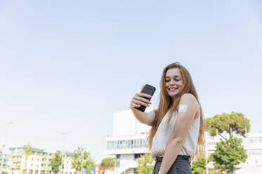 Smiling businesswoman taking selfie through mobile phone after taking vaccination in front of sky - EIF01484