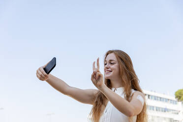 Smiling businesswoman gesturing while taking selfie through mobile phone in front of sky - EIF01487