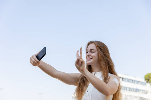 Lächelnde Geschäftsfrau gestikuliert, während sie ein Selfie mit ihrem Mobiltelefon vor dem Himmel macht - EIF01487