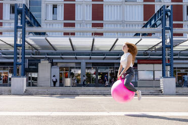 Unbeschwerte weibliche Freiberuflerin springt mit Hüpfball bei Gebäude während sonnigen Tag - EIF01503