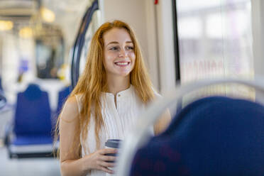 Lächelnde Freiberuflerin mit langen Haaren, die eine Einweg-Kaffeetasse hält und im Zug wegschaut - EIF01521