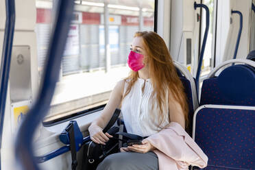 Businesswoman in protective face mask looking through window while commuting in train - EIF01522
