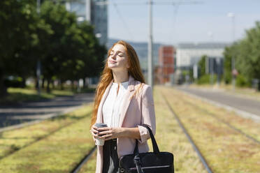 Redhead businesswoman with eyes closed standing at railroad track during sunny day - EIF01527